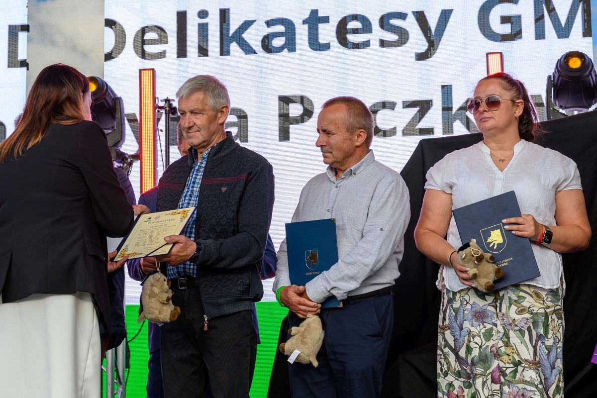 Na zdjęciu troje ludzi otrzymuje nagrody na scenie. Mają ozdobne dyplomy i pluszowe maskotki w rękach. Tło to jasny ekran z tekstem.