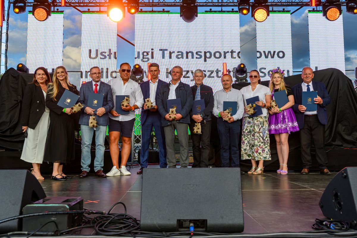 Grupa ludzi stoi na scenie z dyplomami i statuetkami, ubrana elegancko. Tło ozdobione kolorowymi światłami. Za nimi widoczne są trzy duże ekrany.