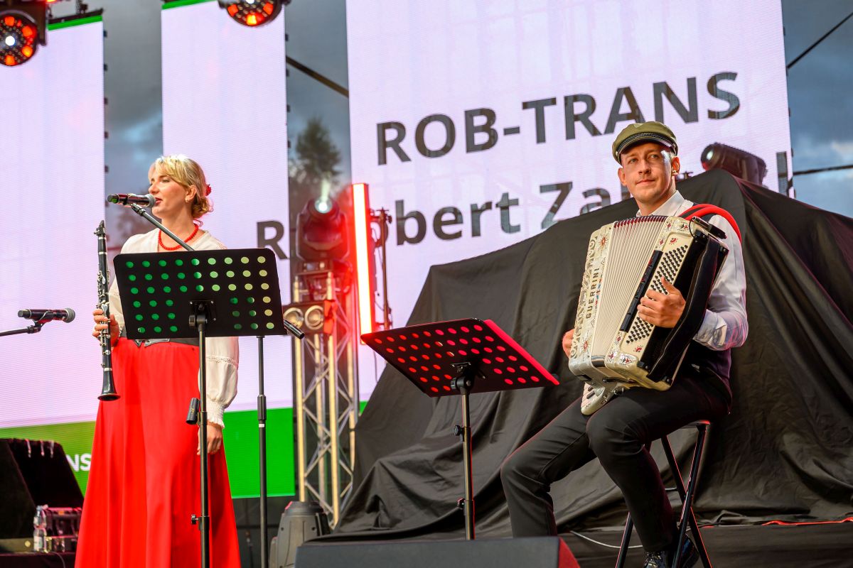 Na zdjęciu jest duet muzyczny: kobieta gra na klarnecie, mężczyzna na akordeonie. Scena jest oświetlona ciepłym światłem, w tle duży ekran z napisem. Dominują kolory czerwony i czarny.
