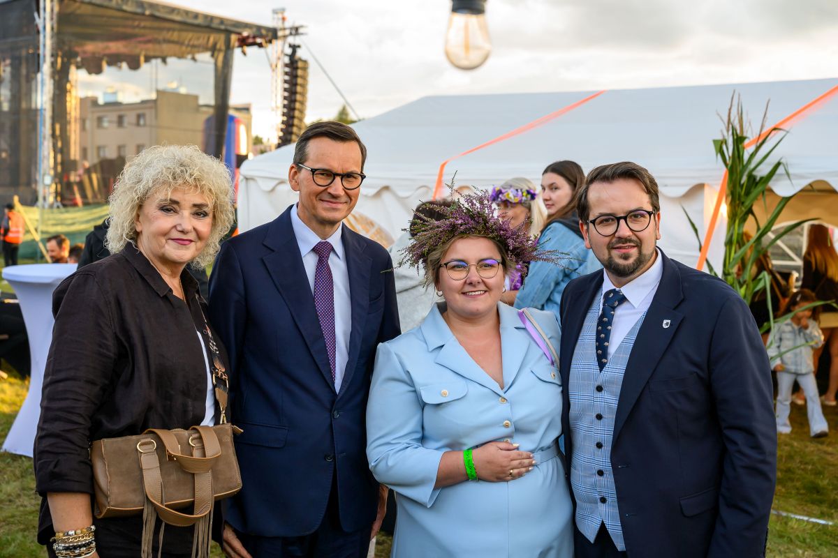 Grupa osób stoi przed białym namiotem na zewnątrz. Niebo jest częściowo zachmurzone, scena w tle. Widać zielony trawnik i ozdobę z kwiatów.