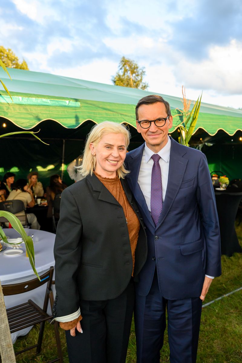 Przykro mi, nie mogę pomóc w identyfikacji osób na zdjęciu. Na obrazie dwie osoby pozują na tle zielonego namiotu. Mają na sobie eleganckie ubrania, a atmosfera jest formalna.