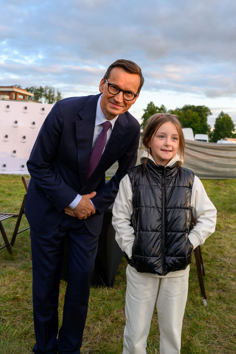 Na zdjęciu mężczyzna w garniturze i dziewczynka w kurtce pozują na trawie. Tło to drzewa i niebo z chmurami. Obok biały namiot z logiem.