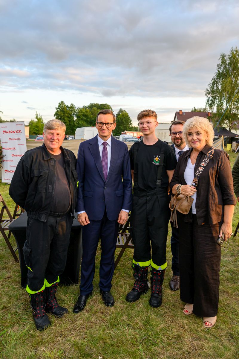 Na zdjęciu grupa osób stoi na trawie. Niebo jest zachmurzone, w tle widać drzewa i zabudowania. Kolory to głównie zieleń, niebieski oraz odcienie brązu.