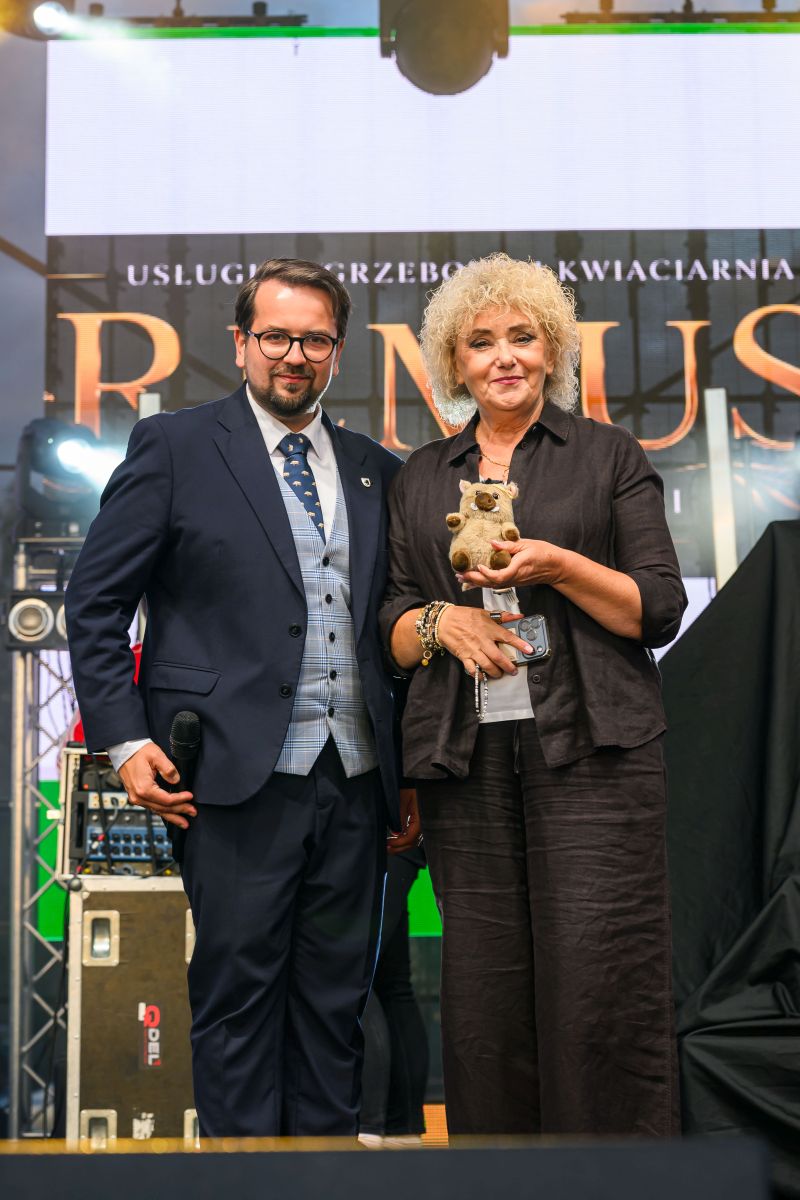 Dwóch ludzi stoi na scenie. Kobieta trzyma pluszaka w kolorze brązowym. Tło zawiera duże litery i oświetlenie sceniczne.