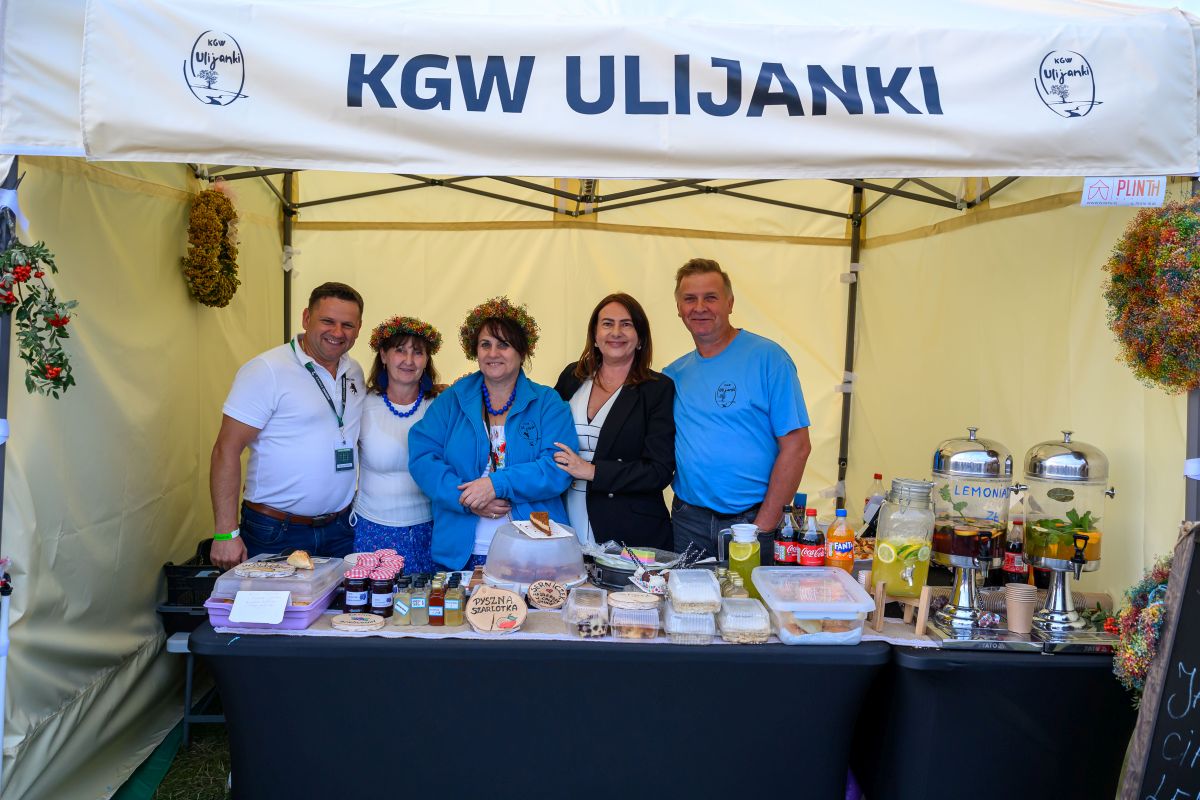 Grupa ludzi stoi pod białym namiotem z napisem KGW Ulijanki. Na stole są przetwory, soki i ciasta. Tło kremowe, dekoracje z wieńcami.