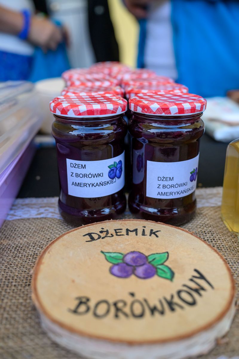 Słoiki dżemu z borówki amerykańskiej z czerwonymi pokrywkami stoją na stole. Na drewnianym krążku widnieje napis Dżemik Borówkowy. Tło rozmyte, dominuje niebieski.