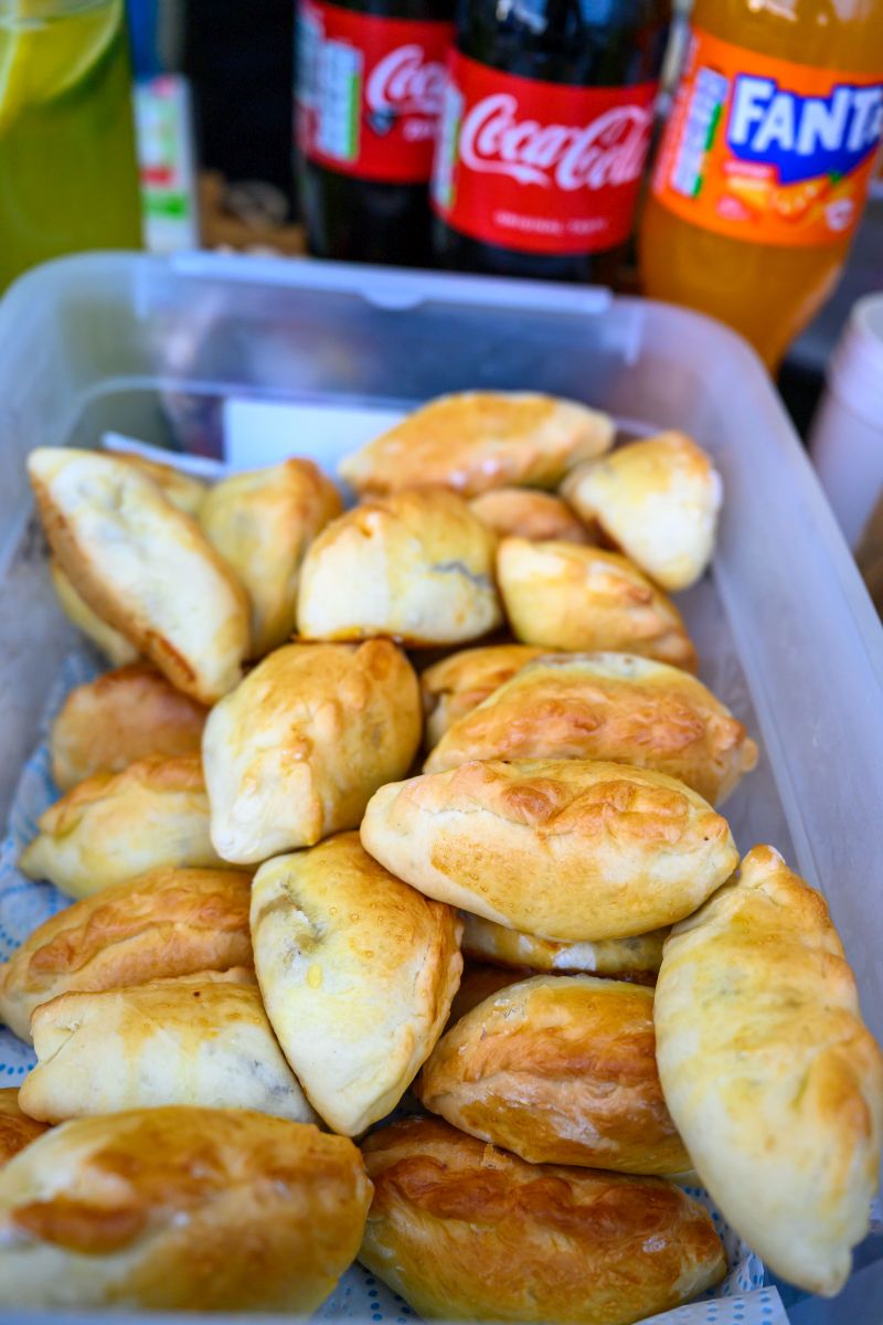 Plastikowe pudełko pełne złocistych empanad. W tle butelki napojów gazowanych. Sceneria kuchenna o żywych kolorach.