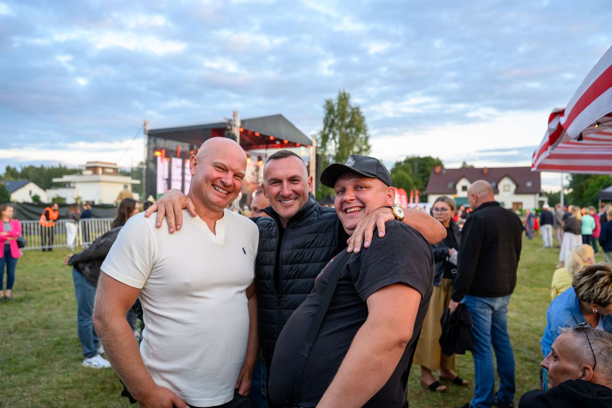 Trzech mężczyzn uśmiecha się na tle sceny plenerowej. Niebo jest zachmurzone, a trawa zielona. W tle widoczni ludzie i domy.