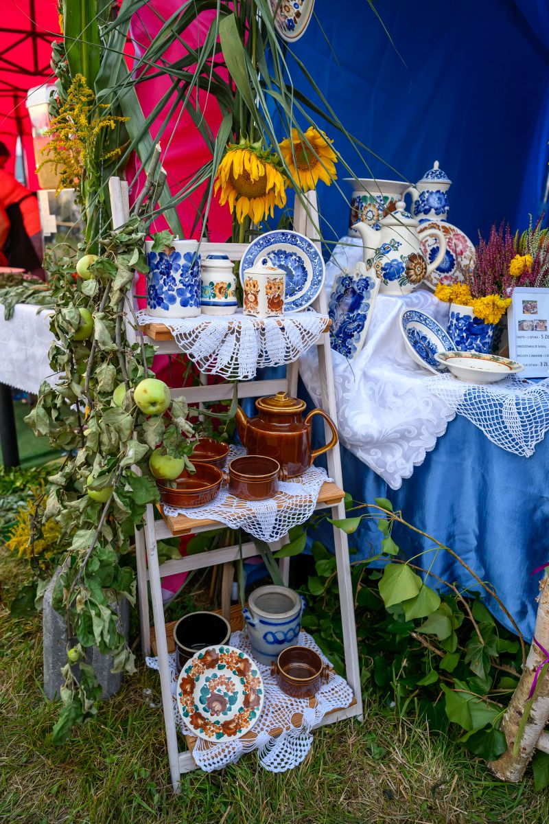 Stojak z ceramiką na tle niebiesko-różowego namiotu. Dekoracje z żółtymi kwiatami i zielonymi liśćmi. Klasyczne naczynia z wzorami.