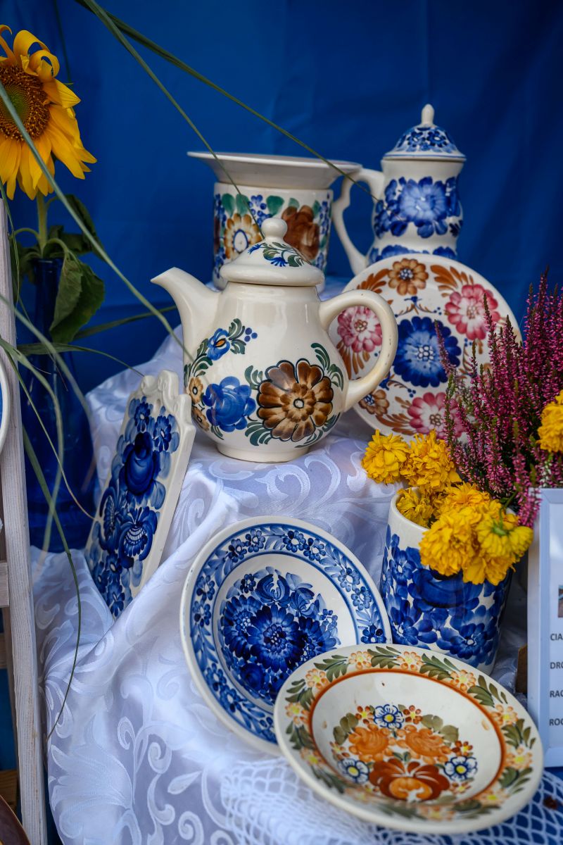 Zestaw porcelany z kwiatowym motywem na niebieskim tle. Dominują kolory: biel, niebieski, brązowy i różowy. Obok wazon z żółtymi kwiatami.