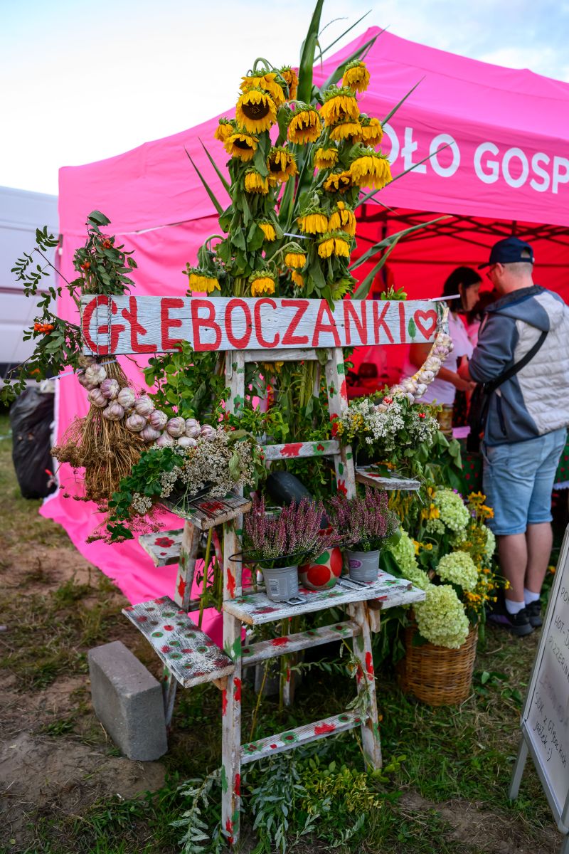 Stoisko z kwiatami i czosnkiem na tle różowego namiotu. Duże drewniane napisy Głęboczanki. Obok ludzie rozmawiający.