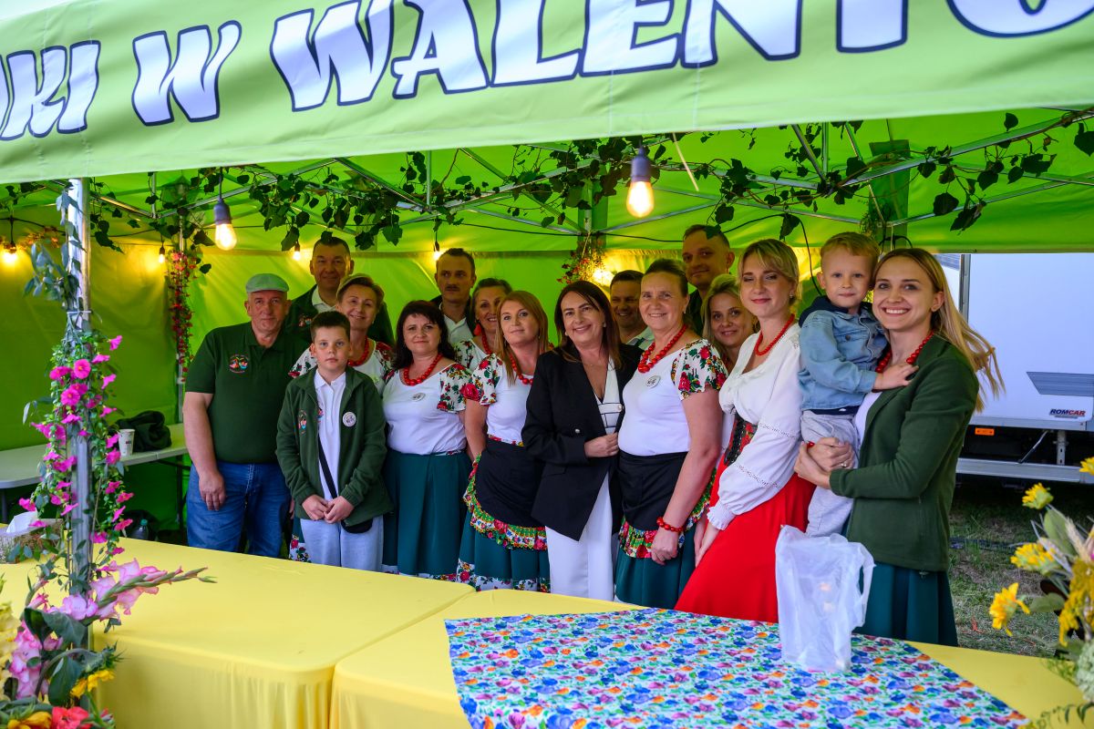 Grupa ludzi pod zielonym namiotem z napisem, ubrana w regionalne stroje. Tło ozdobione kwiatami. Na pierwszym planie żółte stoły.