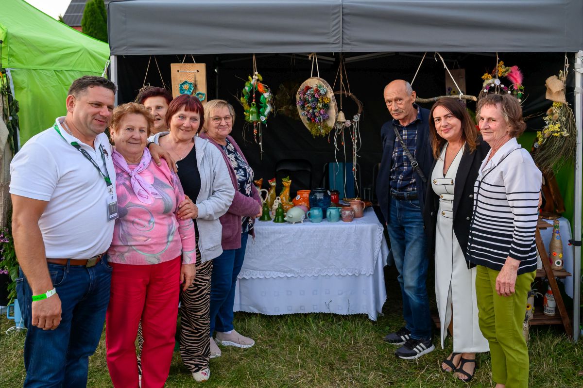 Grupa ludzi stoi przed namiotem z rękodziełem. Tło to zielony i szary namiot z kolorowymi dekoracjami i ceramiką. Wszyscy się uśmiechają.