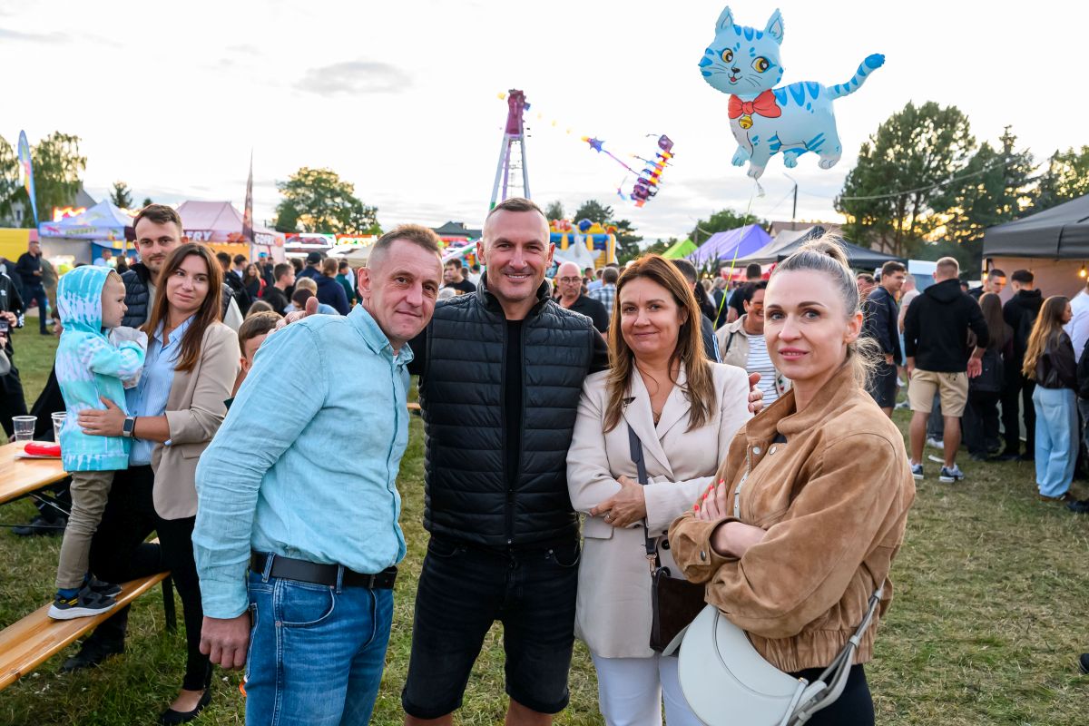 Grupa ludzi na festynie w parku. Kolorowy balon w kształcie kota unosi się w powietrzu. W tle wesołe miasteczko i jasne niebo.