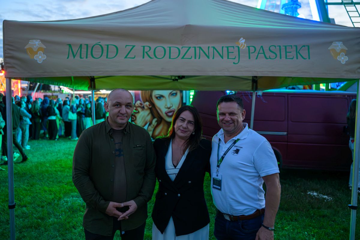 Trzy osoby stoją pod beżowym namiotem z napisem Miód z rodzinnej pasieki. W tle zielone i niebieskie światła oraz czerwony van. Trawa na pierwszym planie, a za osobami tłum ludzi.