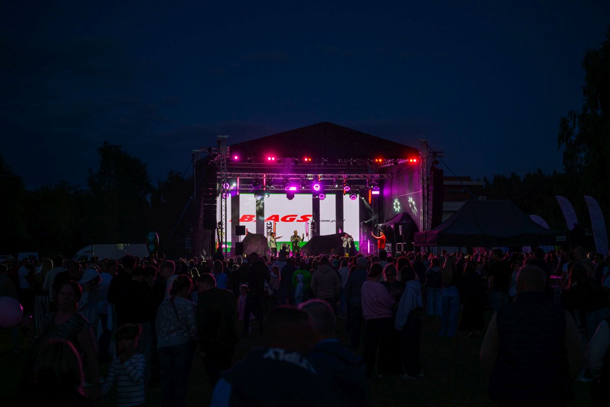 Nocny koncert na scenie z fioletowymi światłami. Tłum widzów z przodu, drzewa w tle. Wyświetlacz z napisem na scenie.