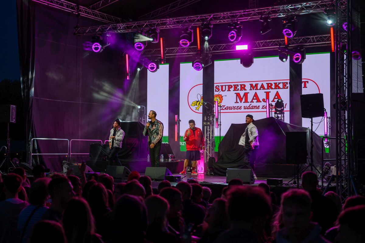 Na scenie występują artyści przed publicznością. Tło jest ciemne z kolorowymi światłami i reklamą supermarketu. Atmosfera jest żywa i dynamiczna.