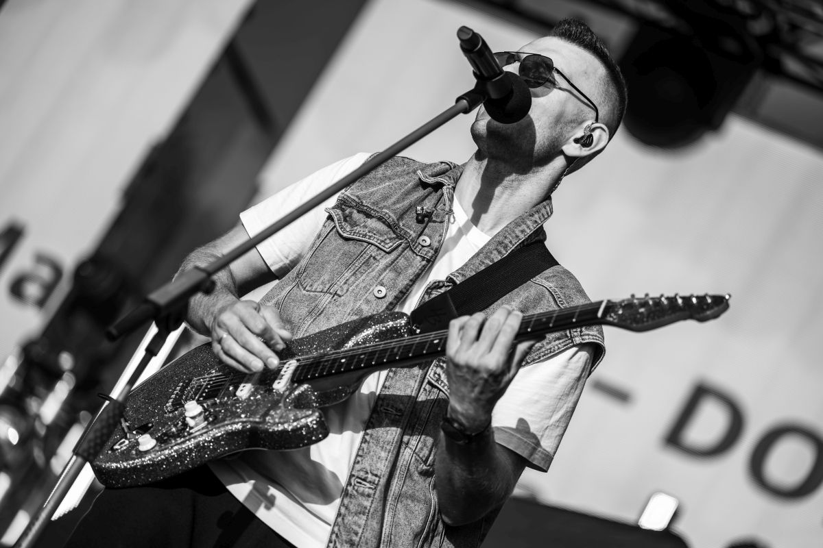 Muzyk w okularach i kamizelce gra na gitarze elektrycznej, śpiewając do mikrofonu. Czarnobiałe zdjęcie na scenie. Tło rozmyte, jasne.