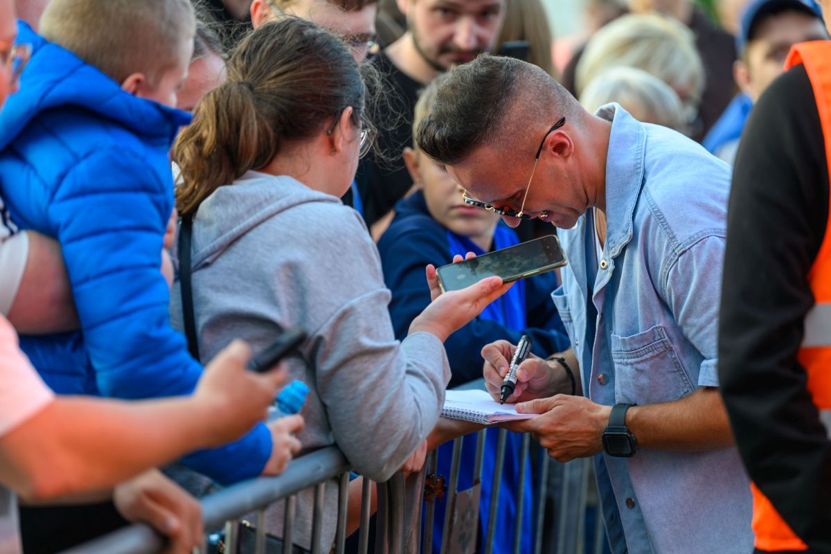 Mężczyzna w okularach podpisuje autograf przy barierce. Obok niego stoi osoba w szarej bluzie, trzymająca telefon. Tłum w tle, dominujące kolory to niebieski i szary.