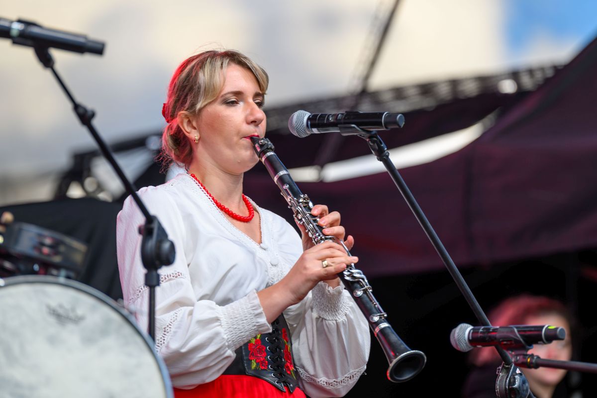 Kobieta w białej bluzce i czerwonej spódnicy gra na klarnecie na scenie. Tło jest ciemne, z mikrofonem z przodu. Widać detale instrumentu.