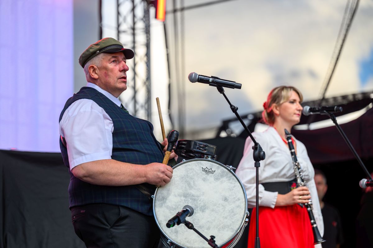 Dwóch muzyków występuje na scenie; mężczyzna w kapeluszu gra na bębnie, kobieta w czerwonej spódnicy gra na klarnecie. Scena jest na zewnątrz, pod niebieskim niebem. Mikrofony stojące przed nimi podkreślają atmosferę koncertową.