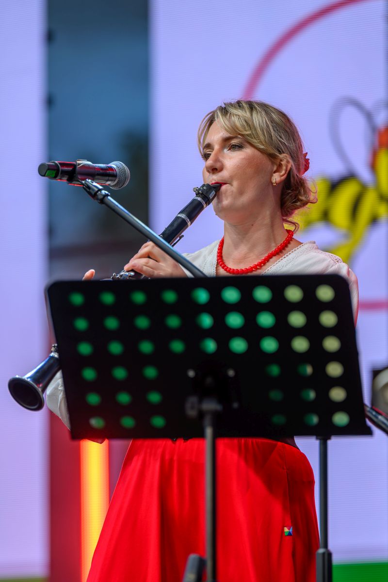 Osoba gra na klarnecie, ubrana w białą bluzkę i czerwoną spódnicę. Tło jest kolorowe, z mikrofonem obok. Przed nią stoi pulpit nutowy.