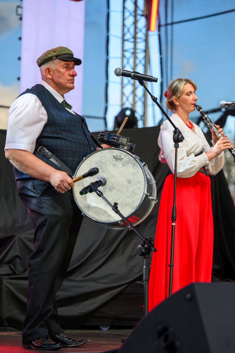 Dwóch muzyków na scenie, mężczyzna gra na bębnie, a kobieta na klarnet. Kobieta ma czerwoną spódnicę, mężczyzna jasną koszulę z kamizelką. Tło zewnętrzne, jasne niebo i scena oświetlona światłami.
