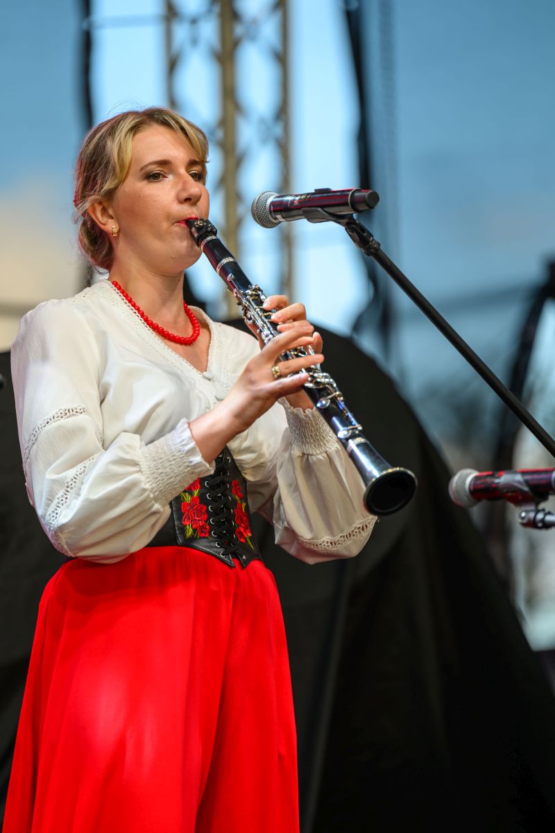 Kobieta w stroju ludowym gra na klarnecie, ubrana w białą bluzkę i czerwoną spódnicę. W tle sceniczne oświetlenie. Instrument zbliżony do mikrofonu.