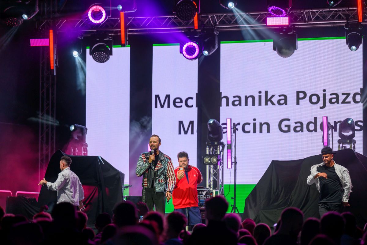 Na scenie trzech mężczyzn, jeden śpiewa. W tle jasne ekrany z tekstem i kolorowe reflektory. Na widowni ciemne sylwetki.