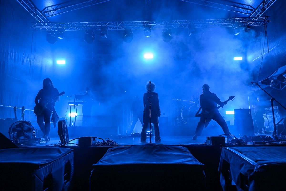 Na scenie w niebieskim świetle występuje zespół muzyczny. Przód zajmuje wokalista, otoczony przez gitarzystów. Tło wypełnia gęsty dym sceniczny.