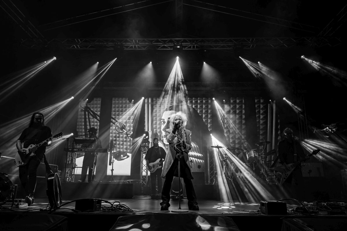 Na zdjęciu jest zespół rockowy na scenie. Dominuje czarno-biały kontrast i intensywne reflektory. W tle widać ekran z dużym wizerunkiem osoby.
