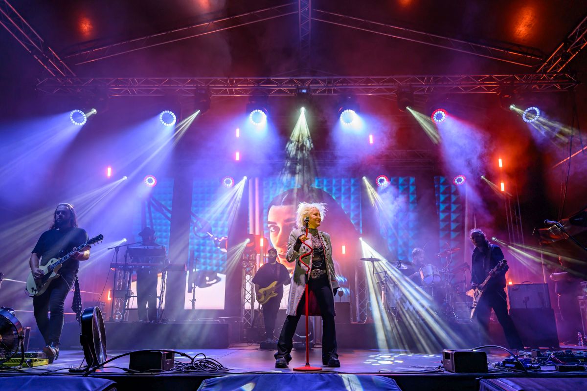 Koncert na scenie z zespołem, dymem i kolorowymi reflektorami w tle. Przeważają kolory fioletowy i niebieski. Wokalistka stoi na środku z mikrofonem.