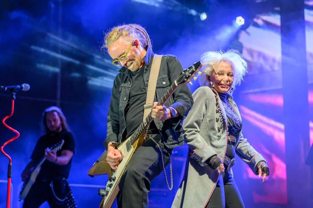 Na scenie para muzyków gra na gitarach. Tło jest fioletowe z jasnymi światłami. Artyści noszą ciemne ubrania i wykazują energię.