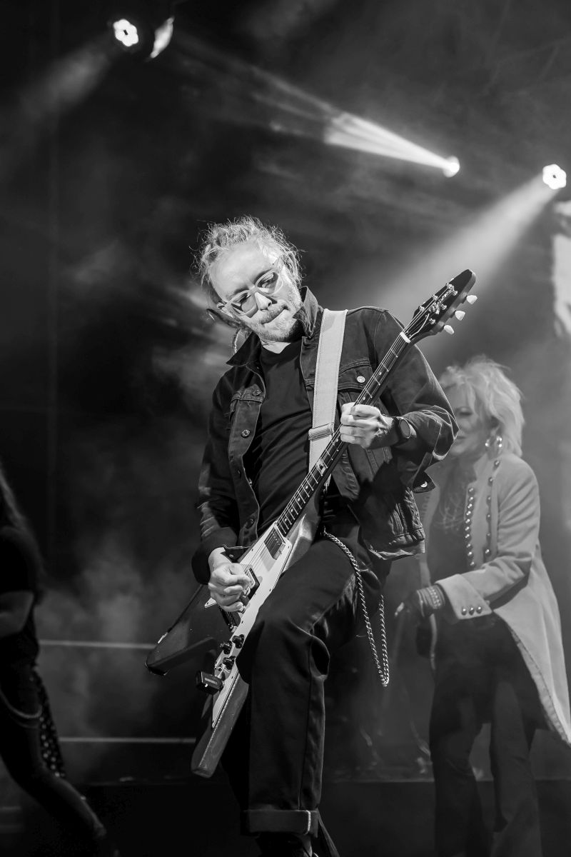 Mężczyzna gra na gitarze elektrycznej na scenie, oświetlony reflektorami. Tło jest ciemne z efektami dymu. Zdjęcie wykonane w czerni i bieli.