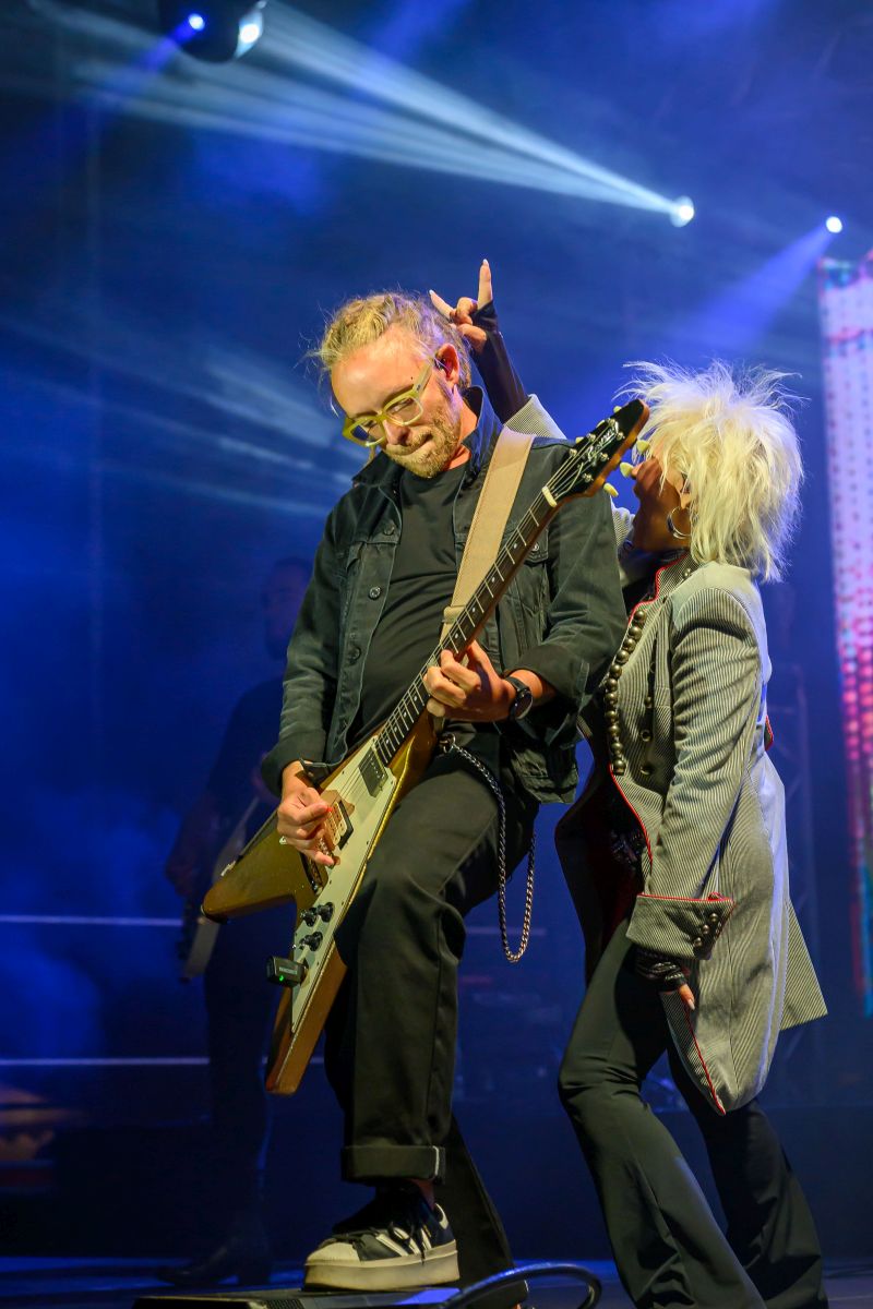 Mężczyzna gra na gitarze na scenie, ubrany na czarno. Obok niego kobieta w szarym płaszczu z białymi włosami. Tło jest ciemne z niebieskimi światłami.