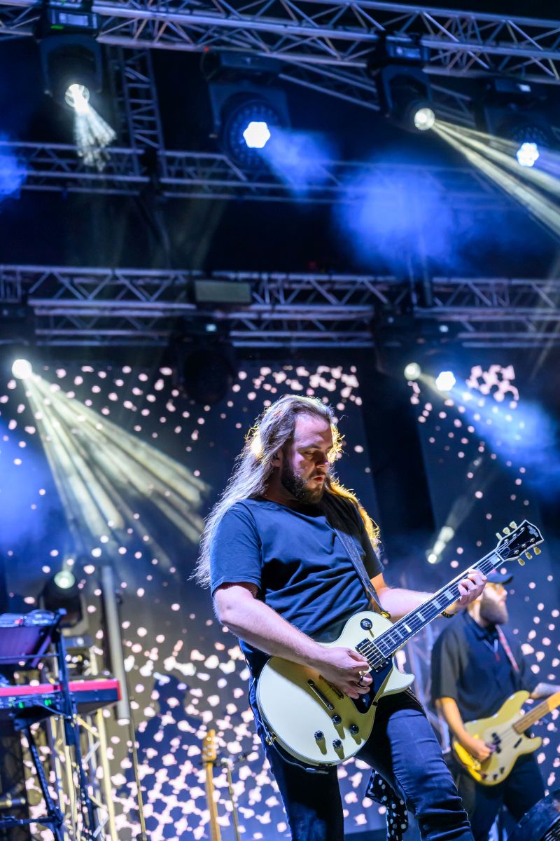 Muzyk gra na gitarze elektrycznej na scenie. Oświetlenie w tle jest kolorowe i dynamiczne. Scena jest pełna reflektorów i dymu.