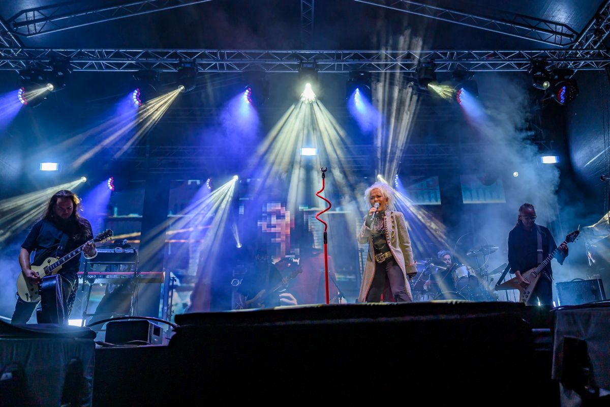 Zespół rockowy na scenie, kolorowe światła i dymy. Centralna postać śpiewa, otoczona gitarzystami. Tło jest ciemne.