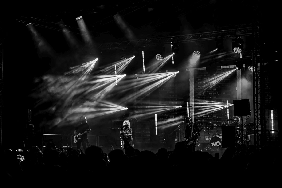 Koncert na scenie z efektami świetlnymi, zespół gra na żywo. Tło ciemne, oświetlenie dynamiczne. Publiczność widoczna w dole zdjęcia.