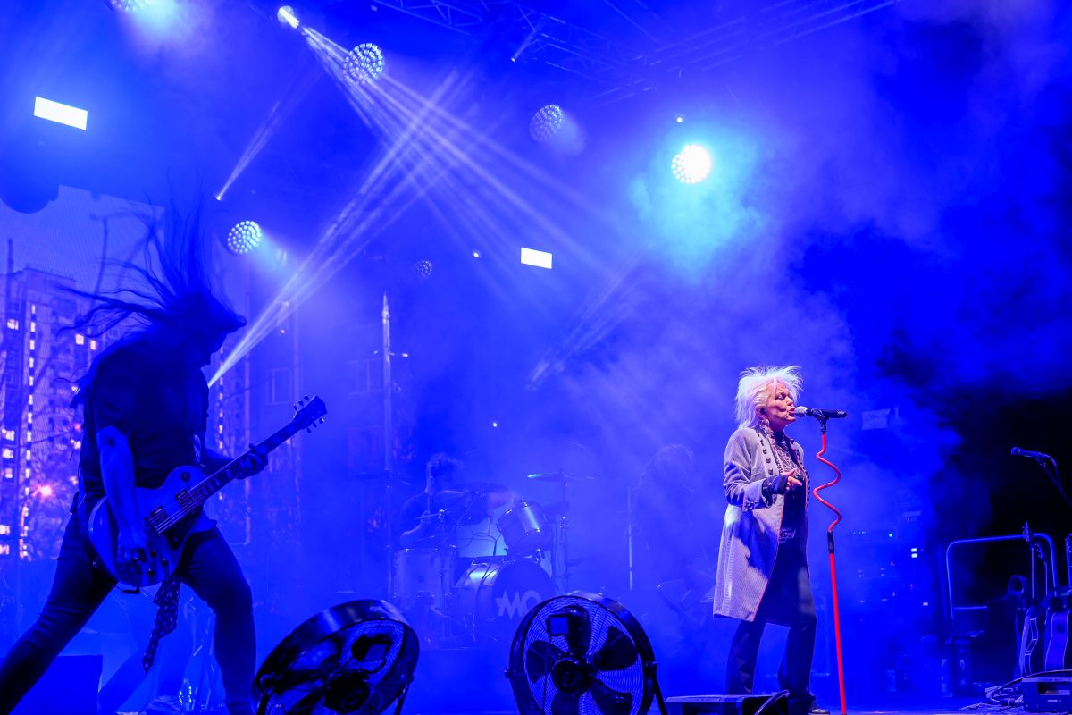 Koncert rockowy z gitarzystą i wokalistką na scenie. Dominują niebieskie światła i dym, tworzące dynamiczną atmosferę. W tle perkusja i intensywne oświetlenie.