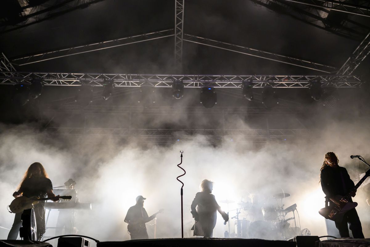 Koncert rockowy na scenie, muzycy w dymie podświetlonym białym światłem, metalowa konstrukcja w tle.