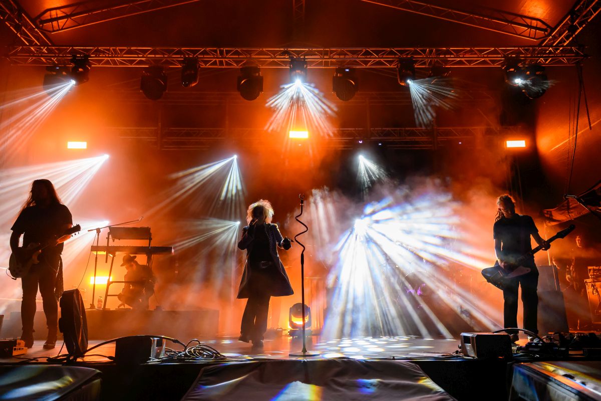 Koncert na scenie z muzykami wśród kolorowych świateł. Dominują pomarańczowe i białe reflektory. Dym na tle dodaje dynamiki.