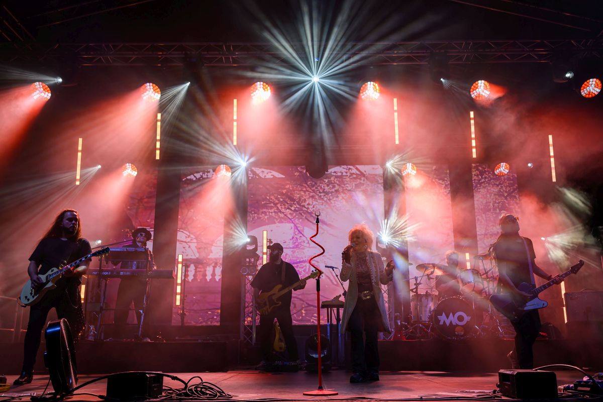 Koncert zespołu na scenie oświetlonej czerwonymi i białymi reflektorami. Dym unosi się w tle, tworząc dynamiczną atmosferę. Muzycy grają na gitarach i instrumentach elektrycznych.