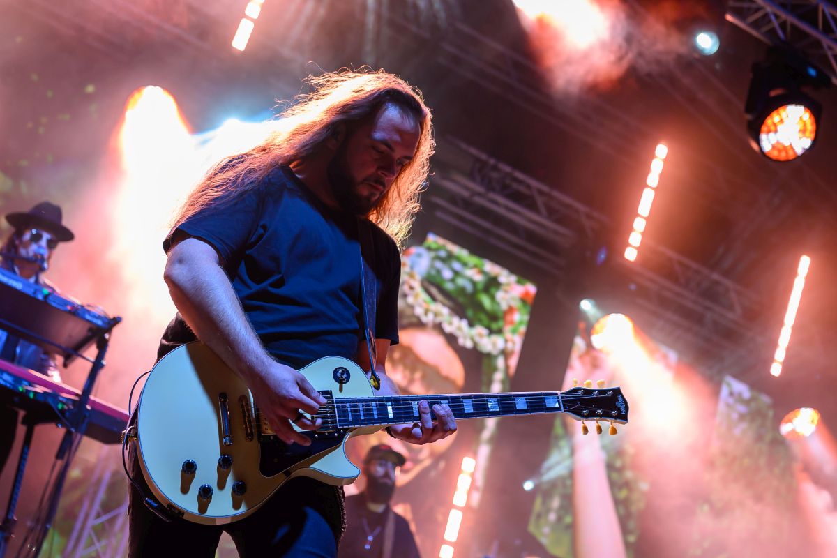 Muzyk z gitarą elektryczną na scenie, otoczony kolorowymi światłami. W tle ekran i aparatura. Scena w dynamicznych barwach.