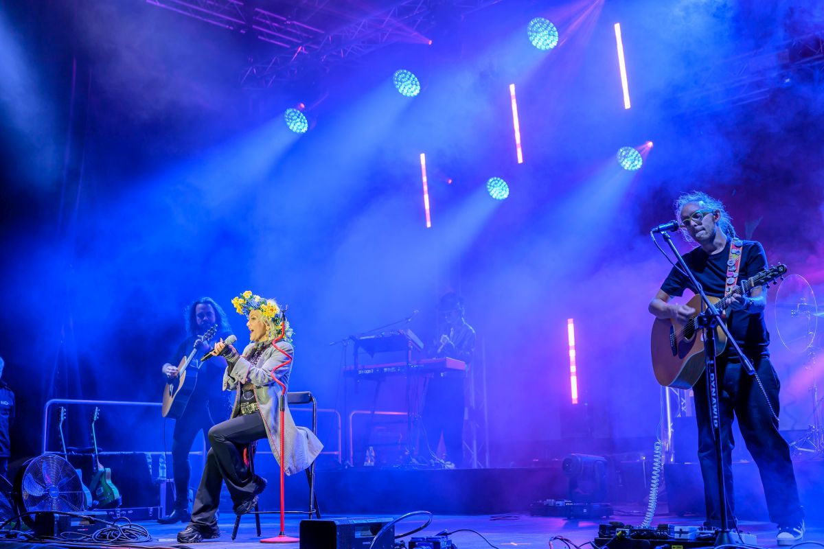 Na scenie artysta z kwiatowym wieńcem śpiewa. Neonowe światła w odcieniach niebieskich i fioletowych tworzą efekt tła. Muzycy grają na gitarach przy jego boku.