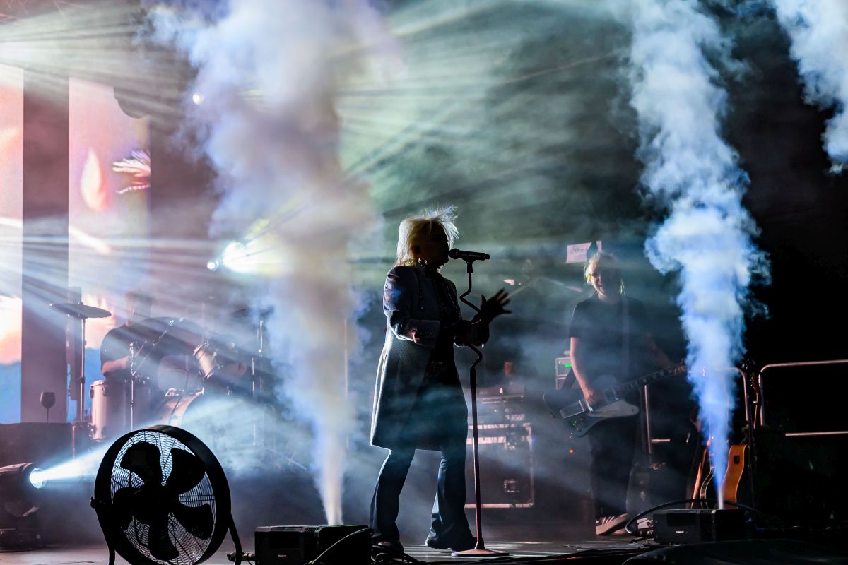 Na scenie widoczny jest wokalista z mikrofonem i gitarzysta. Dym i kolorowe światła tworzą dynamiczny klimat koncertu. Tło mieni się pastelowymi odcieniami.