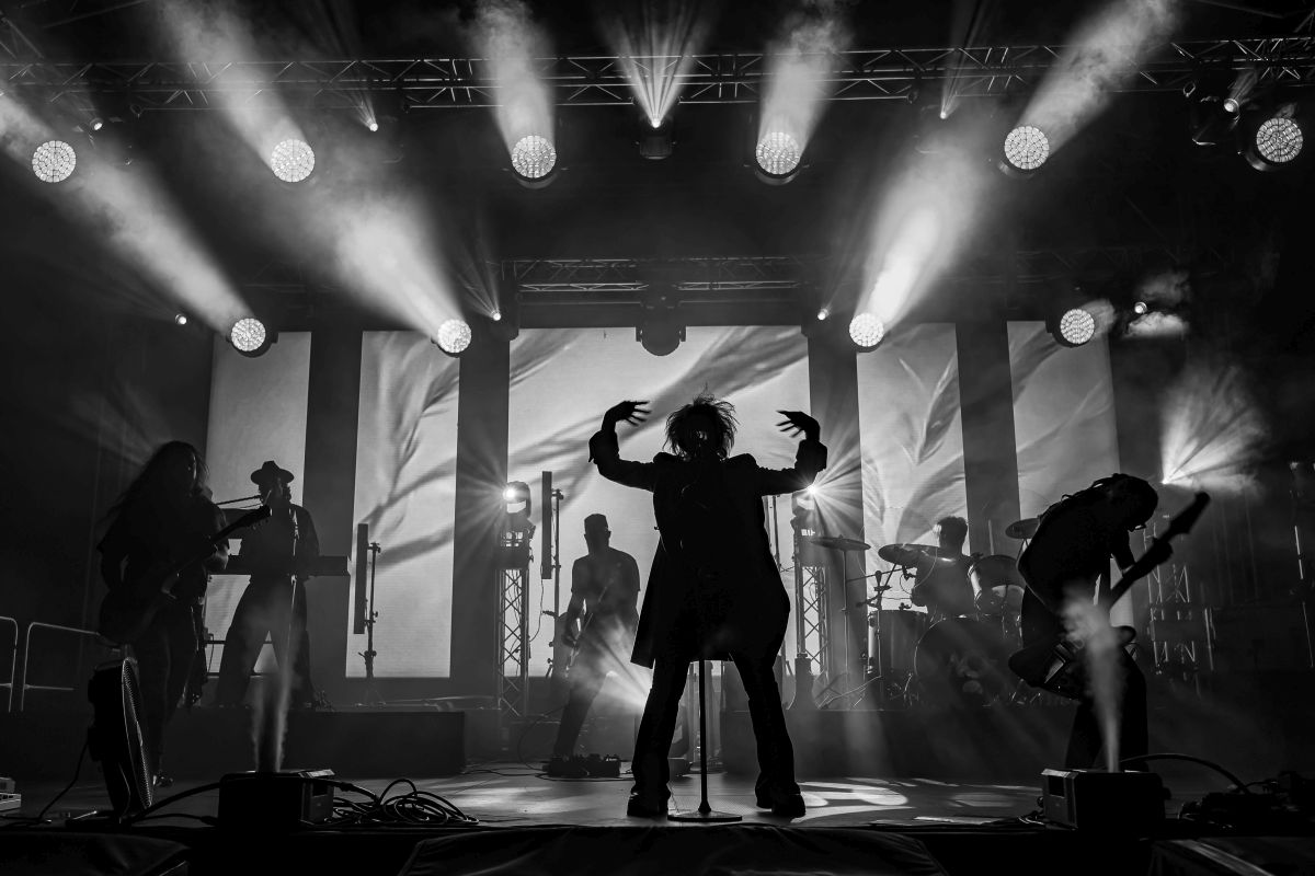 Czarno-biała fotografia przedstawia zespół na scenie w trakcie koncertu. W tle widać jasne światła oświetlające muzyków. Światła tworzą dynamiczną i energiczną atmosferę.
