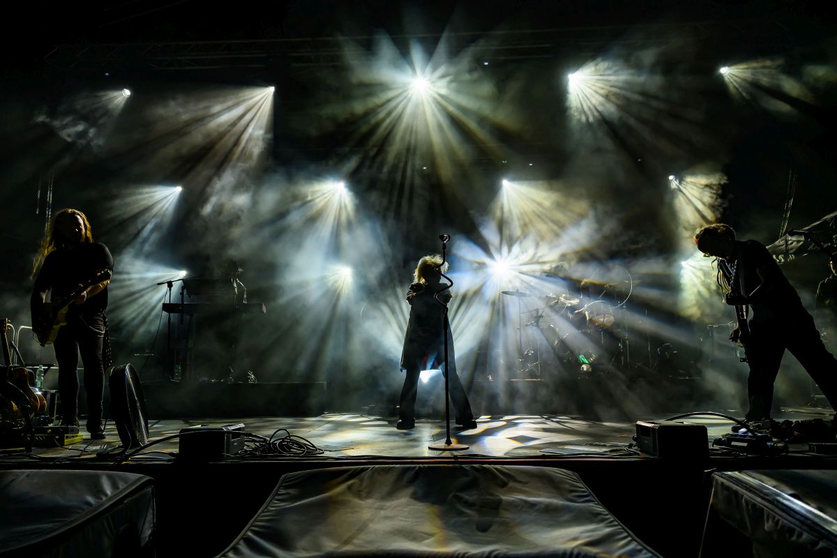 Koncert rockowy na scenie z intensywnym oświetleniem. Światła w różnych kolorach tworzą dynamiczny efekt. Muzycy są w cieniu, a tło jest wypełnione mgłą.