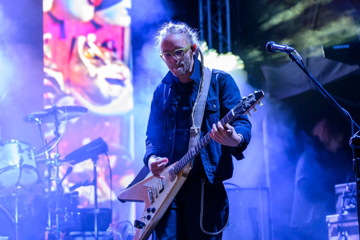 Muzyk gra na gitarze na scenie podczas koncertu. Tło jest kolorowe z intensywnym oświetleniem. Widać instrumenty wśród mgły.
