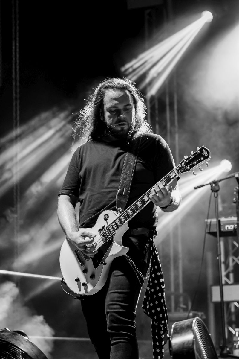 Mężczyzna gra na gitarze elektrycznej na scenie, oświetlony jasnym światłem reflektorów. Tło to dym i sprzęt muzyczny. Zdjęcie jest czarno-białe.