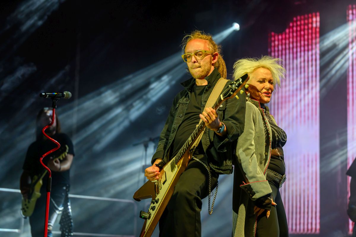 Na zdjęciu duet muzyczny na scenie: gitarzysta i wokalistka. Jasne reflektory oświetlają scenę, a tło jest ciemne. Kolorystyka ciepła, akcenty czerwienia.
